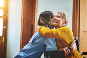 Woman in yellow cardigan smiles while sitting in a wheelchair and hugs a health care worker