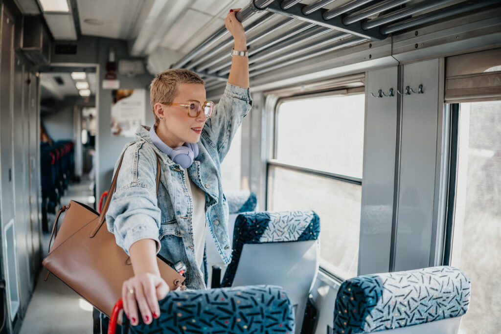 Woman stands inside a train holding on to the overhead rail while carrying a bag and wearing glasses.